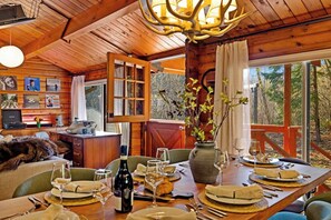 A wooden cabin interior with a dining table set for four, a desk with a computer, and a large window with a view of trees. - Riverfront Log Cabin with Sauna, Hot Tub, Whisky Barn (Rhododendron)