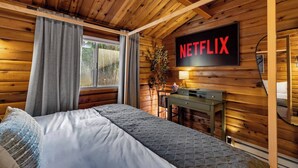 Ein gemütliches Schlafzimmer einer Holzhütte mit einem Bett, einem Schreibtisch und einem an der Wand montierten Fernseher, der 'NETFLIX' anzeigt.