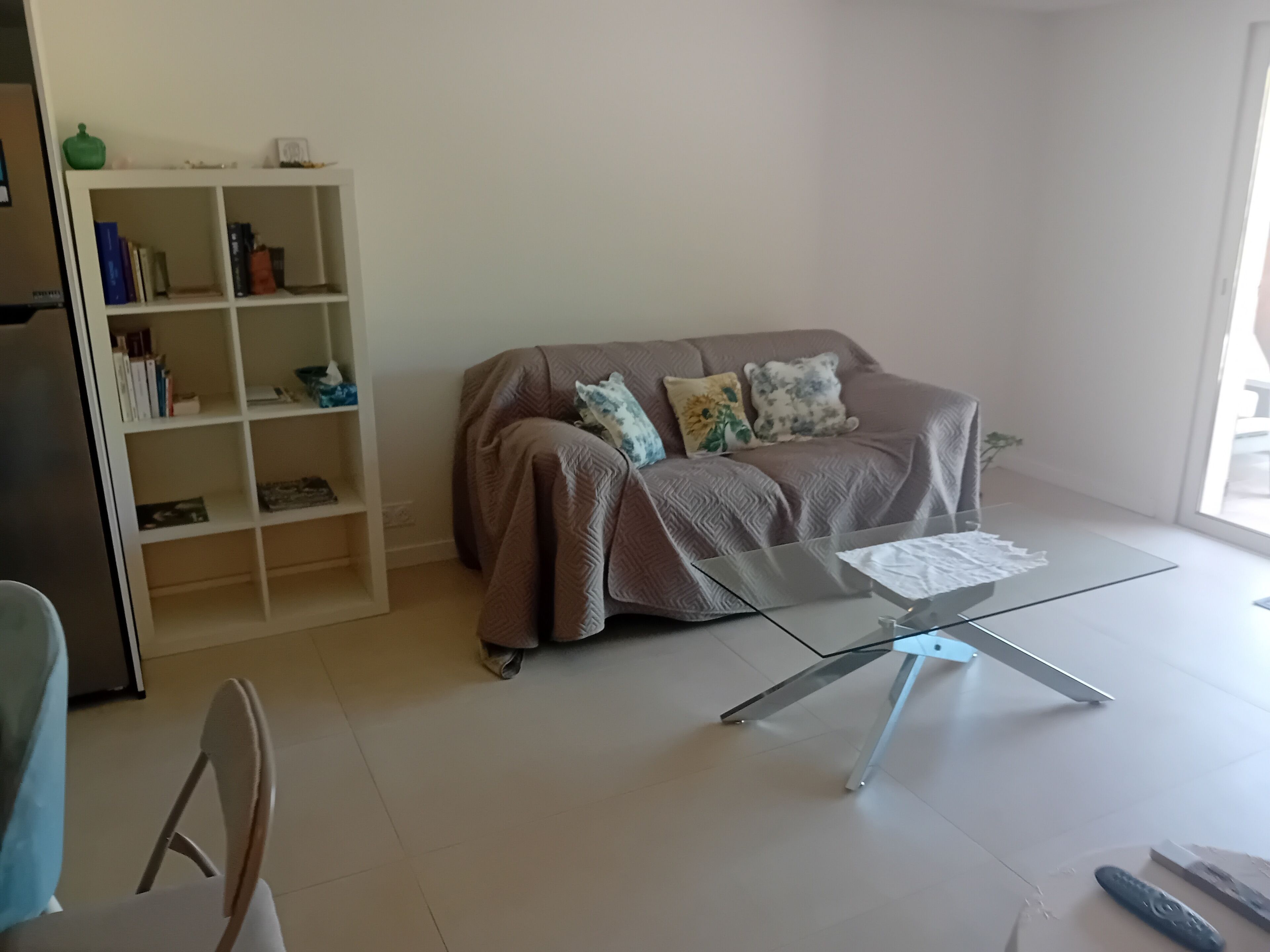 A living room with a glass coffee table, a sofa with patterned cushions, a bookshelf, and a chair.
