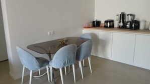Ein Essbereich mit einem Glastisch, vier Stühlen, einem weißen Schrank mit Küchengeräten und einem kleinen dekorativen Gegenstand auf dem Tisch.