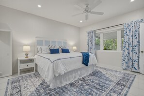 A bedroom with a bed, two bedside tables, a ceiling fan, a window with curtains, and a patterned rug.