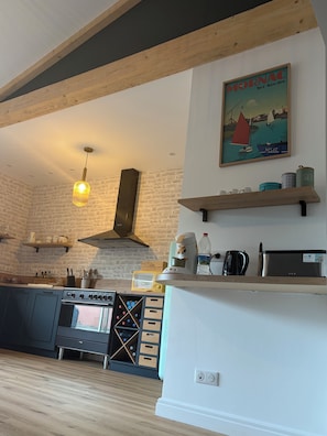 Eine Küche mit Herd, Backofen und Kaffeemaschine, Holzbalken, einer Pendelleuchte und einem Gemälde eines Segelboots an der Wand.