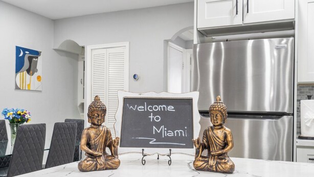 Et køkken med et rustfrit stål køleskab, et tavleskilt med påskriften 'Welcome to Miami', og to gyldne Buddha-statuer på bordet.