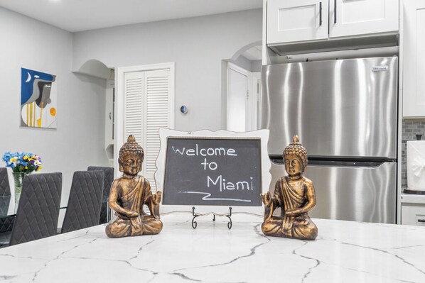 Eine Küche mit einem Edelstahlkühlschrank, einem Kreidetafel-Schild mit der Aufschrift 'Welcome to Miami' und zwei goldenen Buddha-Statuen auf der Arbeitsfläche.