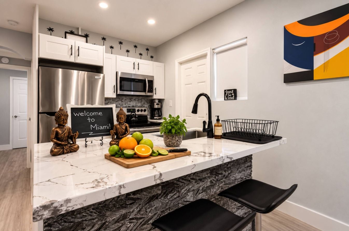 A kitchen with white cabinets, a stainless steel refrigerator, a microwave, and a sink with a black faucet. There is a welcome sign, a Buddha statue, and a cutting board with fruits.
