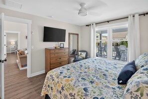 A bedroom with a bed, dresser, TV, and a balcony with chairs. - Second Row Sunset Beach Island West    Coastal Escape 
Luxury Beach House (Sunset Beach)