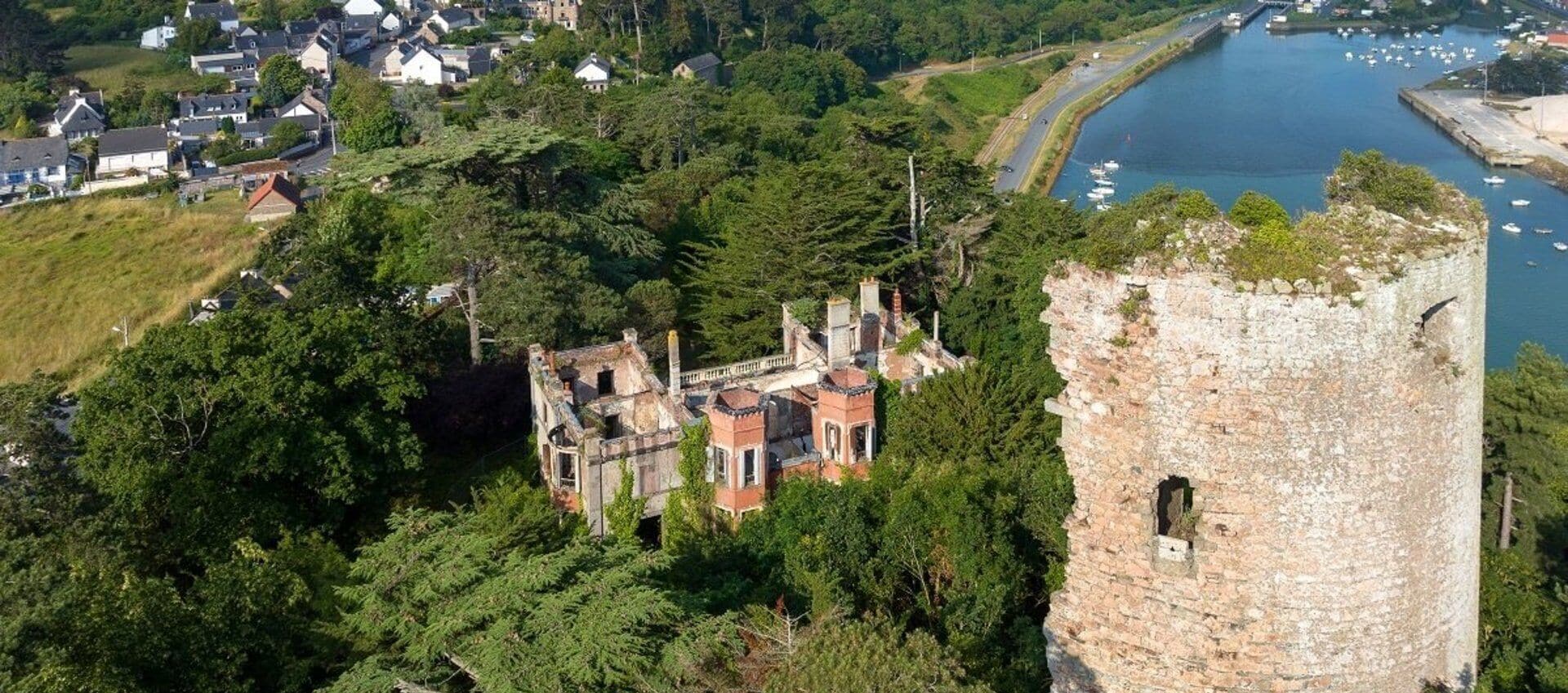 Eine Vogelperspektive eines historischen Gebäudes, umgeben von üppiger Vegetation, mit einem steinernen Turm zur Rechten und einem Gewässer im Hintergrund.