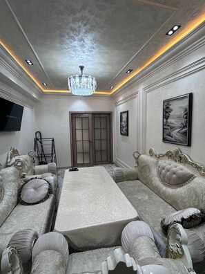 A spacious living room with a large sofa set, a chandelier, and a flat-screen TV mounted on the wall.