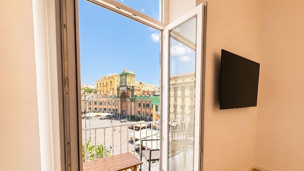 A balcony with a wooden table, a TV mounted on the wall, and a view of a historic building with a clock tower. - Penthouse Napoli Terrace-Dante Rooftop (Naples)