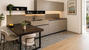 A modern kitchen with a dark countertop, a sink, and cabinets. There is a dining table with two chairs and a vase with flowers. A framed picture of Naples is on the wall. - Penthouse Napoli Terrace-Dante Rooftop (Naples)