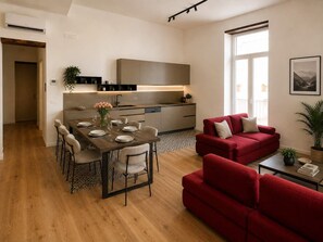 A modern living space with a dining area, a red sofa, and a kitchen with a built-in oven. - Family & Friends Wing at Penthouses Dante. Private wing for groups: 35sqm kitchen/lounge, 3 ensuite bedrooms—30sqm Suite with balcony on Piazza Dante, 20sqm Superior, and a Double room. Access to panoramic rooftop over historic center's ancient domes. (Naples)