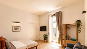 Ein Schlafzimmer mit einem Bett, einem Sofa, einem Fernseher, einem Fenster mit Vorhängen, einer Tür und einer kleinen Pflanze.