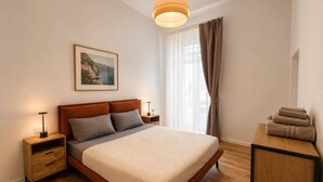 Un dormitorio con cama, mesitas de noche, una lámpara, un cuadro enmarcado, una ventana con cortinas y un escritorio de madera con toallas.