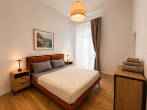 A bedroom with a bed, bedside tables, a lamp, a framed picture, a window with curtains, and a wooden desk with towels. - Dante Sky Apt: Flowers Room & Terrace (Naples)