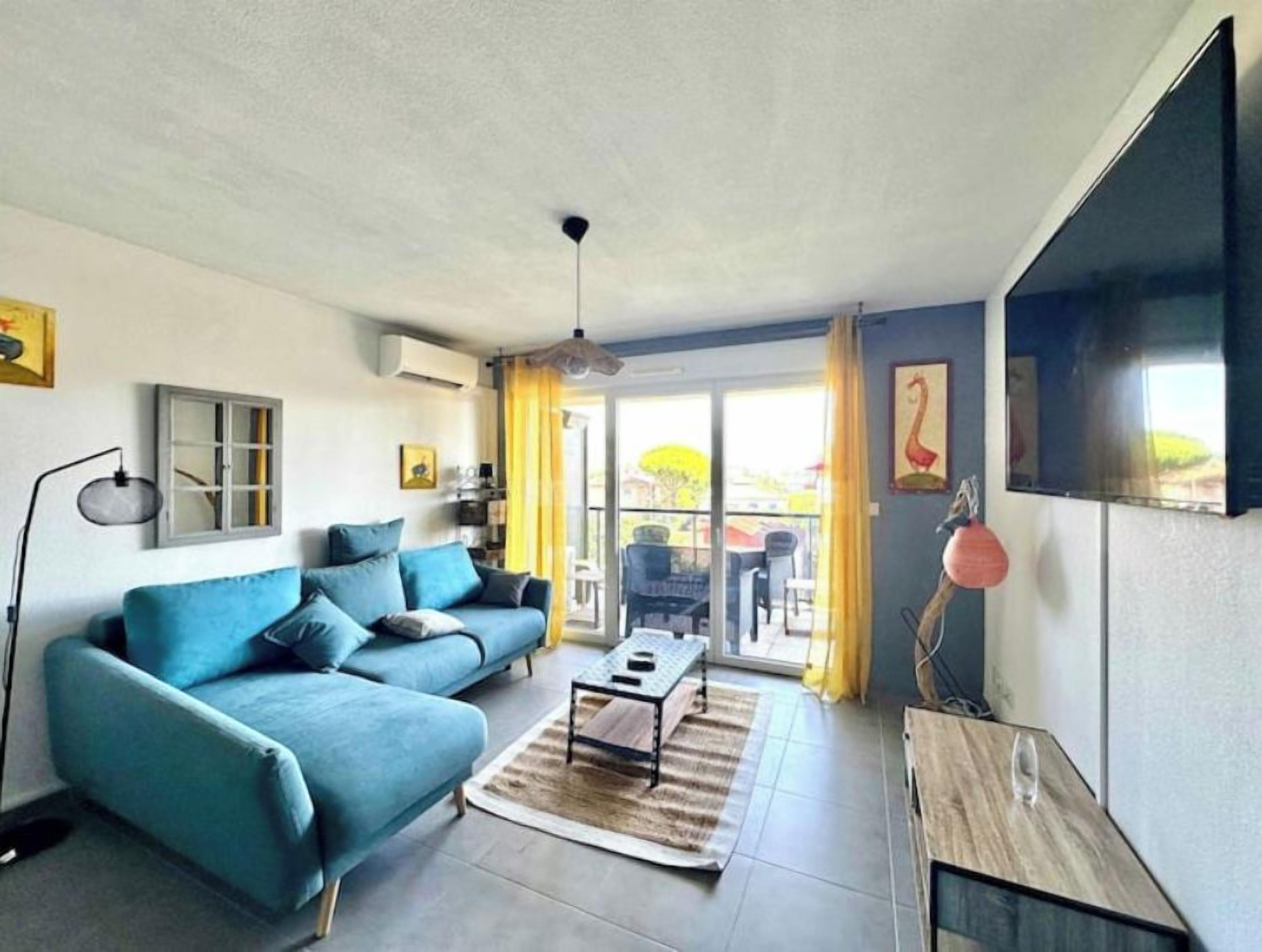A living room with a sofa, a coffee table, and a TV. There is a window with curtains and a door leading outside.