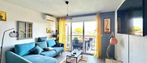 A living room with a sofa, a coffee table, and a TV. There is a window with curtains and a door leading outside.