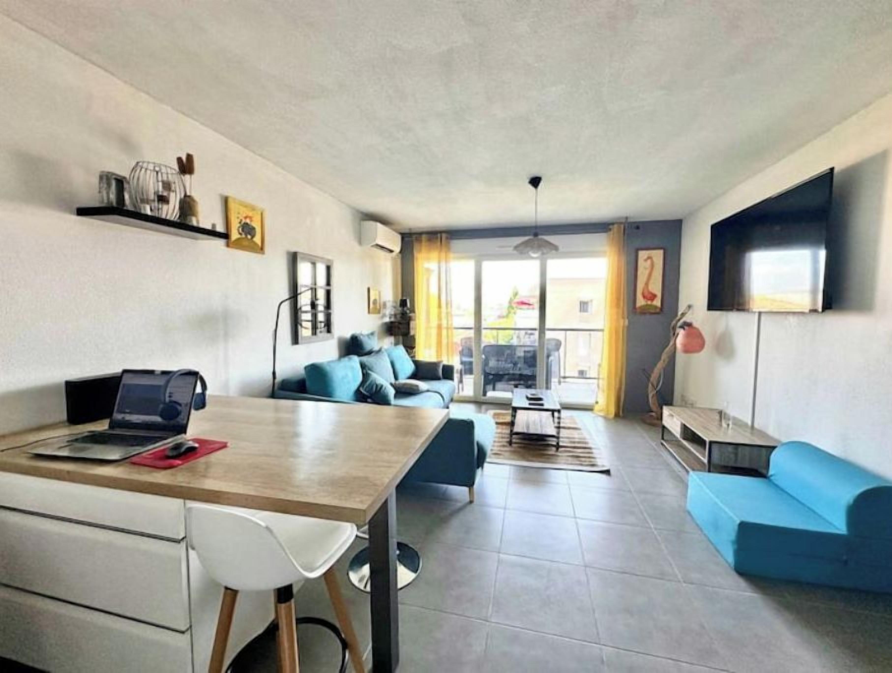 A modern living room with a dining area, a blue sofa, a flat-screen TV, and a laptop on the table.