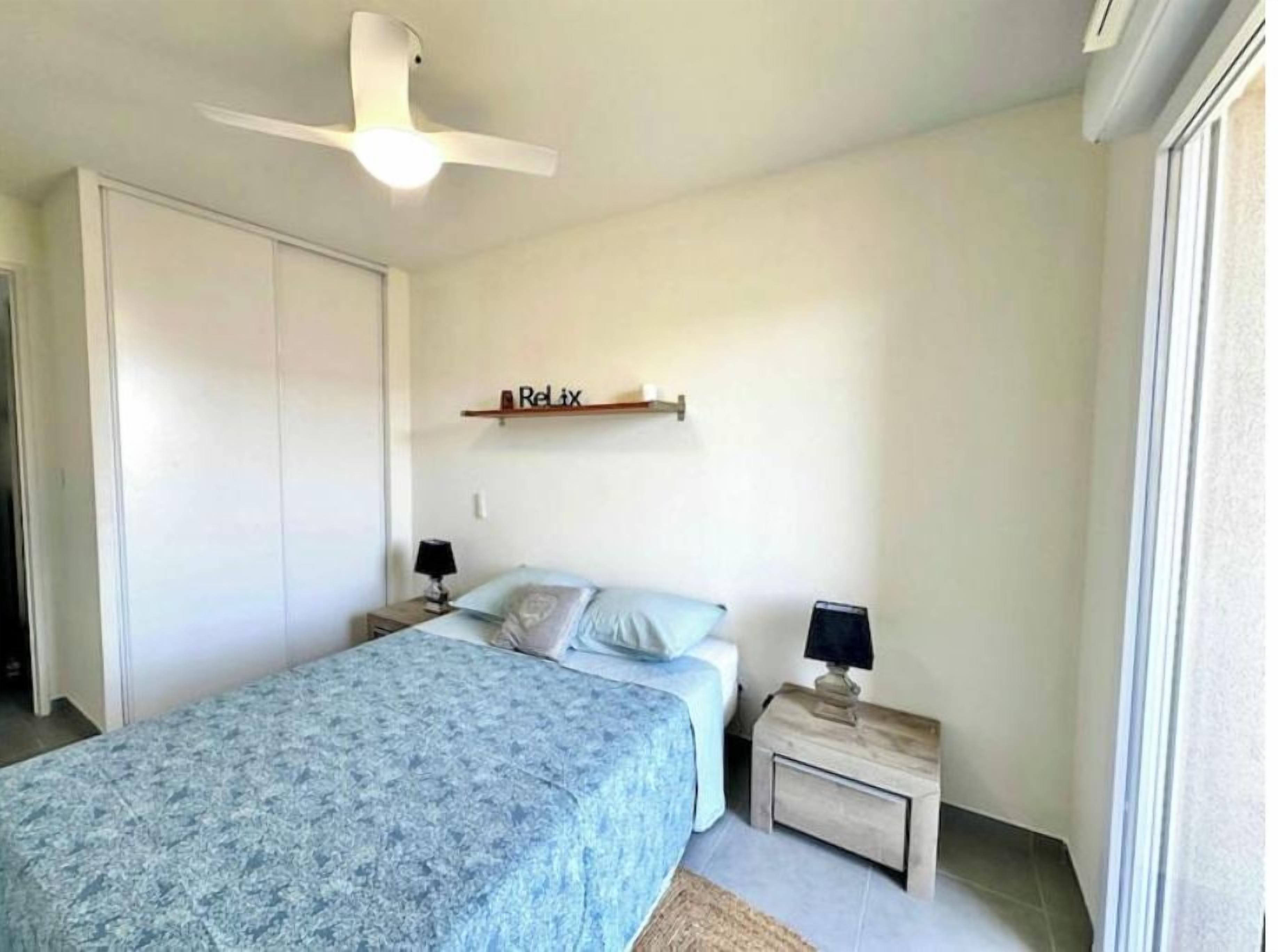 A bedroom with a bed, bedside tables, a ceiling fan, a shelf with a 'Relax' sign, and a window.