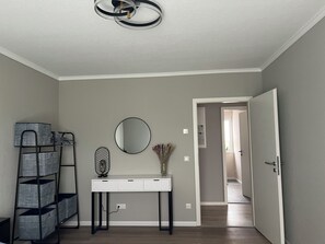 Ein Flur mit einem weißen Konsolentisch, einem runden Spiegel, einem schwarzen Metallregal und einer Tür zu einem anderen Raum.