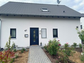 Ein modernes Haus mit einer dunkelblauen Tür, Hausnummer 47, einer Satellitenschüssel und einem kleinen Garten mit Blumen und Pflanzen.