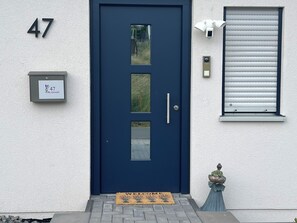 Una puerta azul oscuro con un panel de vidrio, un buzón con el número de la casa, un felpudo y una ventana con persianas.