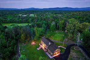 Une grande maison avec un foyer extérieur dans la cour, entourée d’une forêt dense et de collines en arrière-plan.