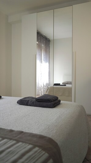 Ein Schlafzimmer mit einem Bett, einem gefalteten grauen Handtuch, einem Kleiderschrank und einem Spiegel, der Bett und Vorhänge reflektiert.