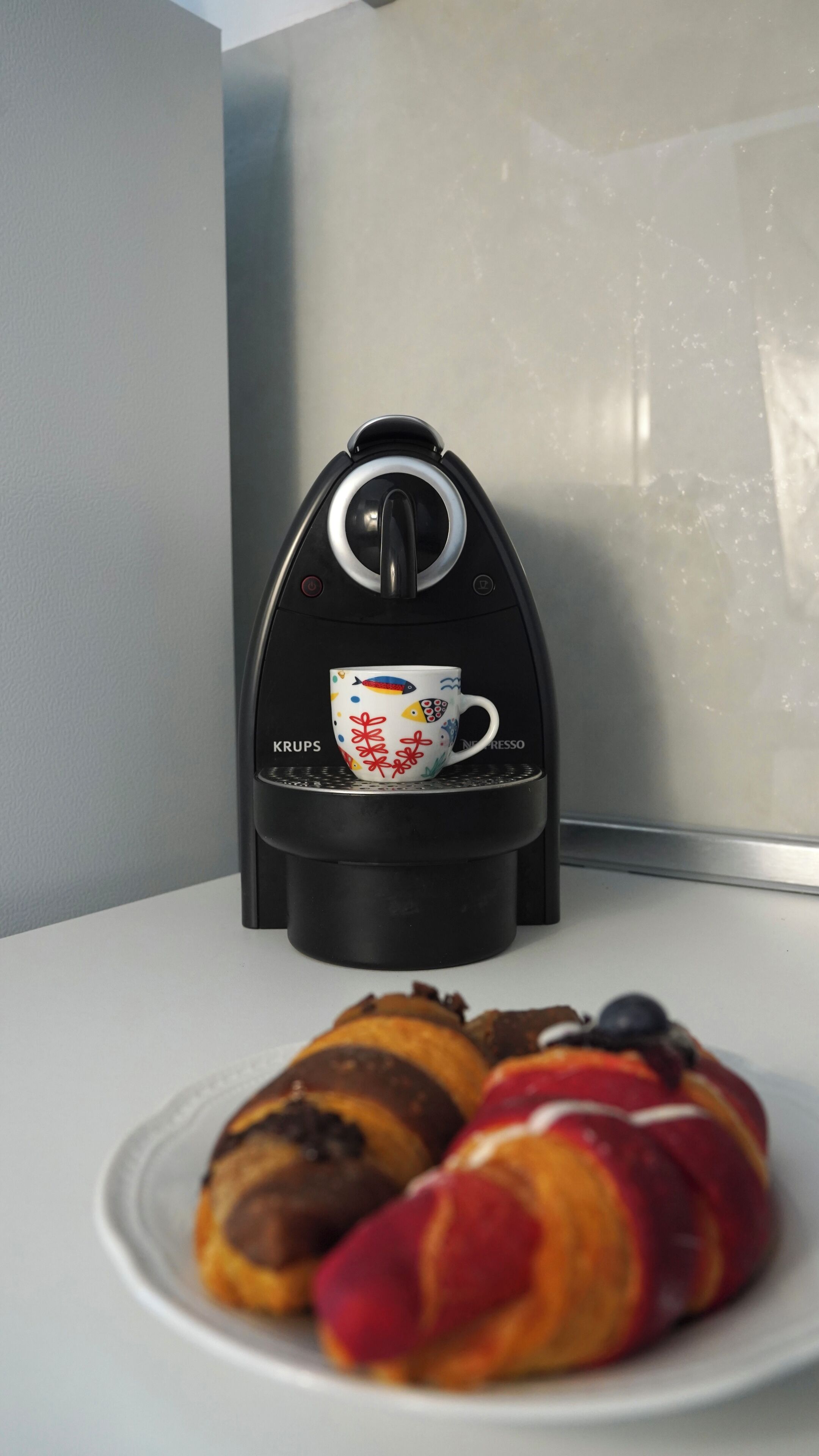 A coffee maker with a cup on its base, a plate with croissants, and a window in the background.