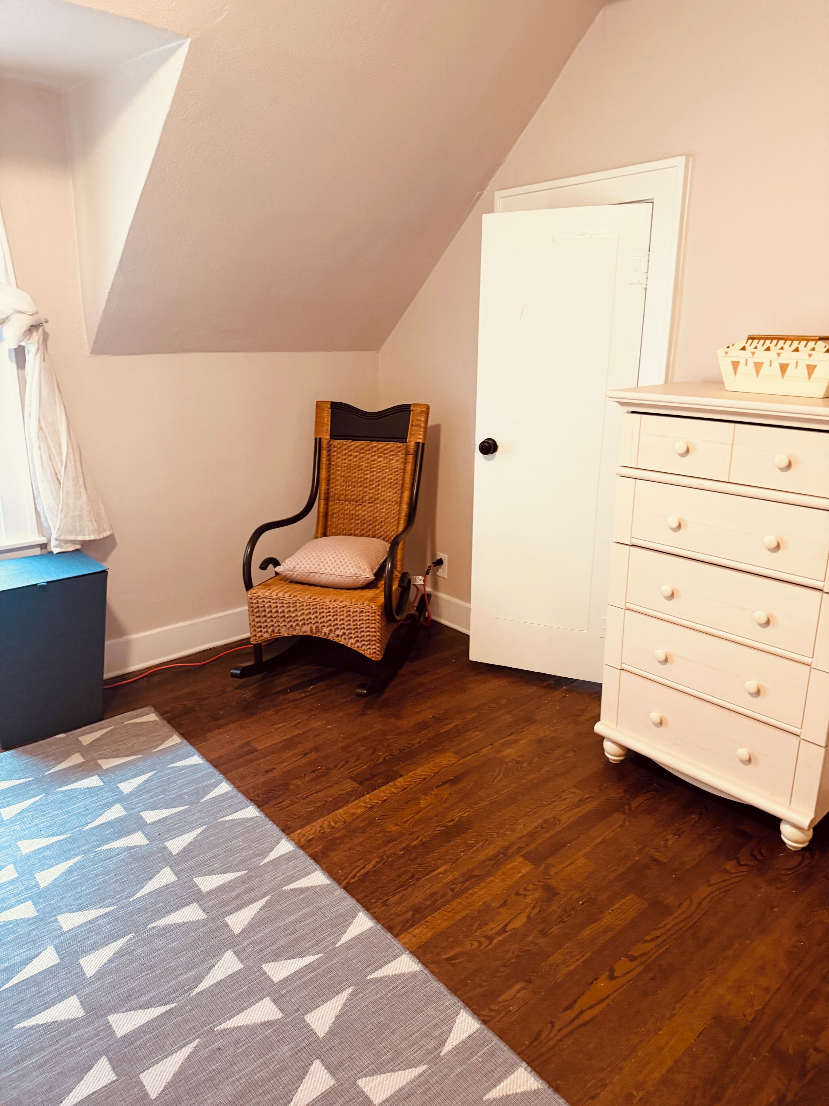 Een kamer met houten vloer, een rieten stoel met kussen, een witte dressoir met lades, en een kamerbreed tapijt met dessin.