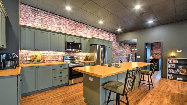 A kitchen with a brick wall, grey cabinets, stainless steel appliances, and a wooden island with bar stools.