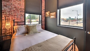 A bedroom with a brick wall, a bed with white bedding, a bedside table with a lamp, and a window with blinds.