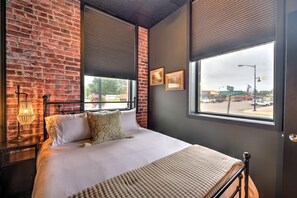 Quarto com parede de tijolos, cama com roupa de cama branca, mesa de cabeceira com abajur e janela com persianas.
