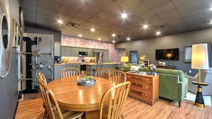 A modern kitchen with a brick backsplash, a dining area with wooden chairs, and a living room with a green sofa and a TV.