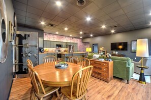 Cozinha moderna com painel de tijolos, área de jantar com cadeiras de madeira e sala de estar com sofá verde e televisão.