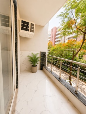 Un balcon orné d’un pot de fleurs, une fenêtre avec des stores et une vue sur un arbre aux fleurs orangées.