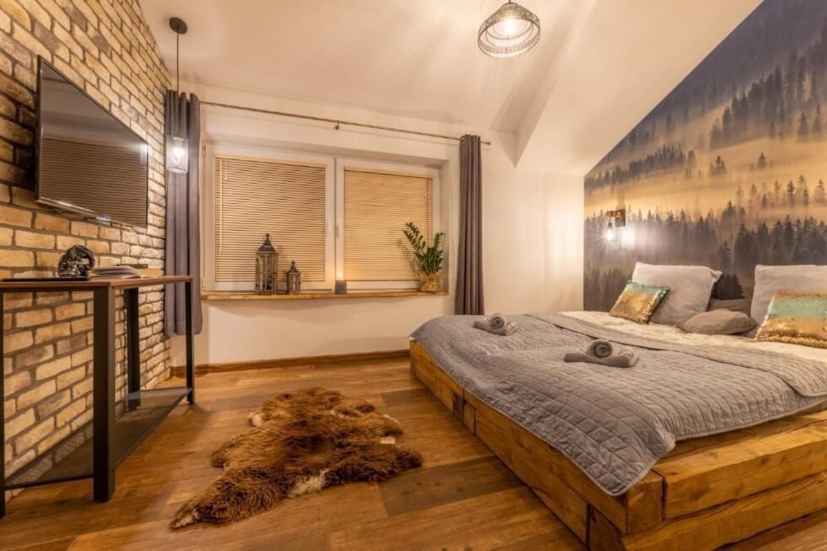A bedroom with a wooden bed, a large window with blinds, a mounted TV, and a forest mural on the wall.