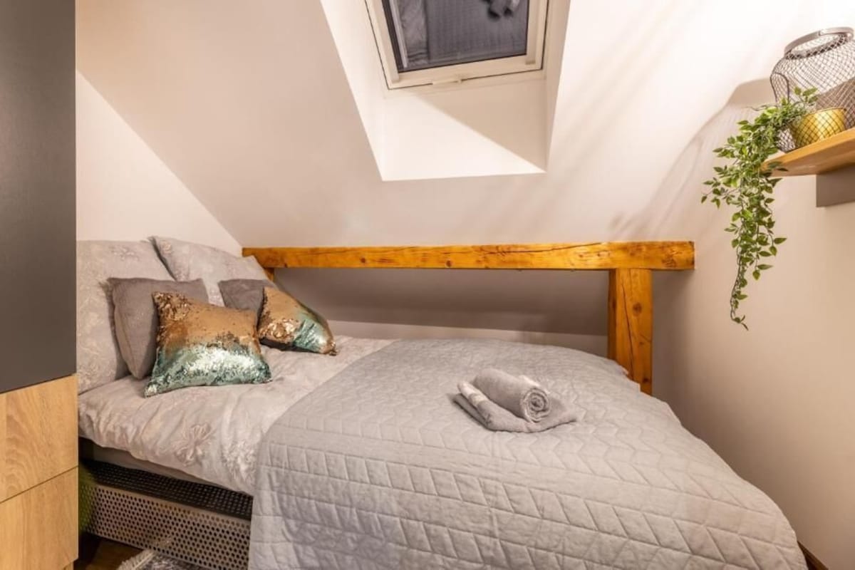 A small, cozy bedroom with a sloped ceiling, a wooden headboard, a bed with a quilted blanket, two decorative pillows, and a small window.