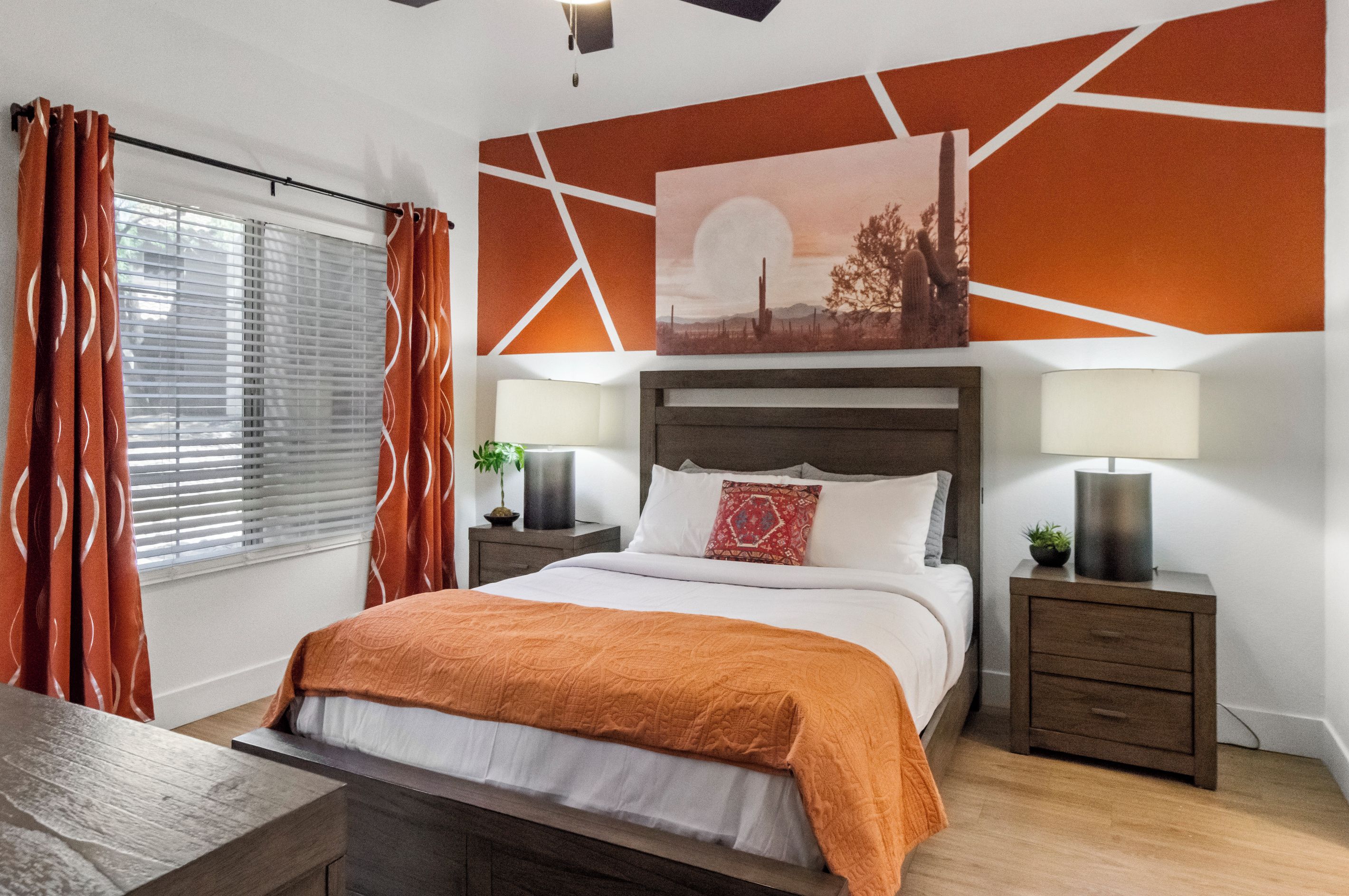 Une chambre moderne avec un lit, deux tables de chevet, une commode, une fenêtre avec des stores et un mur orné d’un motif géométrique et d’un tableau.
