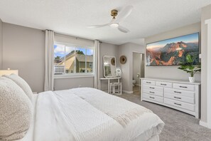 Quarto com cama, penteadeira com espelho, cômoda, ventilador de teto e uma janela grande.