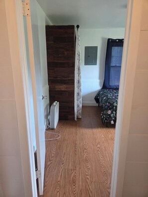 Une pièce avec un plancher en bois, un lit recouvert d’une couvre-lit à motifs floraux, un radiateur blanc et une fenêtre avec un rideau.