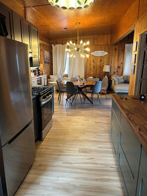 A modern kitchen with a stainless steel refrigerator, wooden cabinets, a dining area with a table and chairs, and a chandelier.