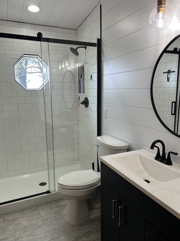 A modern bathroom with a glass shower enclosure, a white toilet, a sink with a black faucet, and a mirror with a round frame.