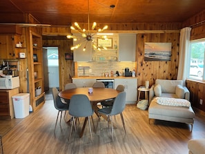 A cozy cabin-style room with a wooden interior, a dining table with chairs, a kitchenette, and a comfortable seating area by the window.