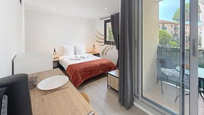 Hotelkamer met een bed, een eettafel, een stoel, een tafeltje met een lamp, een raam met gordijnen, en een balkon met een stoel.