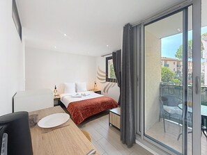 Ein Hotelzimmer mit einem Bett, einem Esstisch, einem Stuhl, einem kleinen Tisch mit Lampe, einem Fenster mit Vorhängen und einem Balkon mit Sitzgelegenheit.