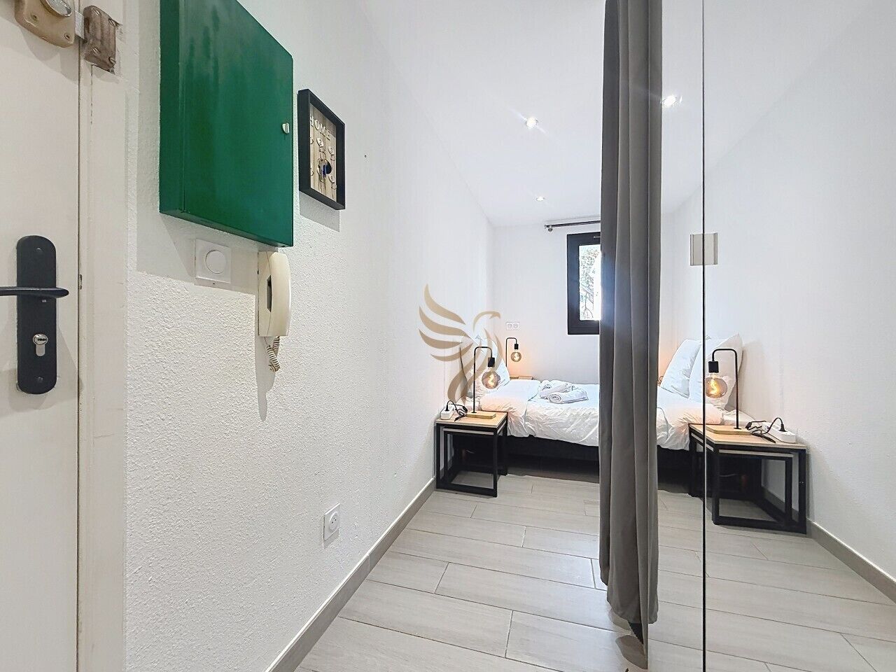 A hotel room with a bed, bedside tables, a green wall-mounted panel, a framed picture, and a door with a lock.