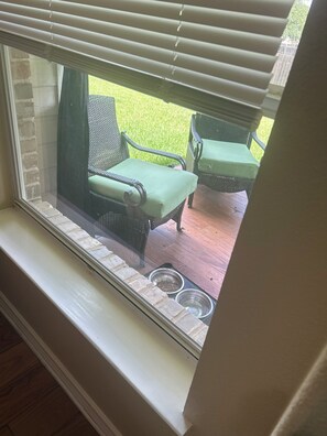 A window with a view of a patio area, including a brick wall, two wicker chairs with green cushions, and two metal bowls on the floor. - Spacious 5-bedroom house in enchanting Austin with AC (Austin)