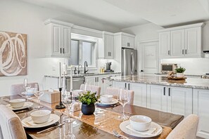 A modern kitchen with white cabinetry, a stainless steel refrigerator, and a dining area set for a meal. - Thalia Bright Designer Home 12 Min To Falls (Thorold)