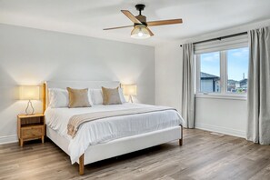 A bedroom with a bed, two bedside lamps, a ceiling fan, a window with curtains, and a view of buildings outside. - Thalia Bright Designer Home 12 Min To Falls (Thorold)