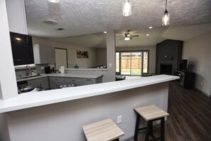 A modern kitchen with a white countertop, bar stools, and a dining area with a fireplace. - Modern Spacious Home in Quiet Neighborhood (La Porte)
